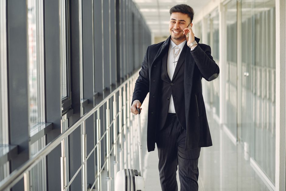 Happy ethnic male in formal suit standing in modern hallway while holding suitcase in hand and talking on mobile phone